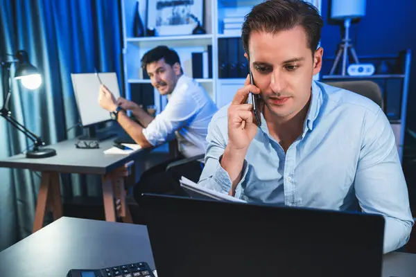 真剣なビジネスマンは夜間に新しいプロジェクトを作成するために顧客と電話をかけ 同僚は机の上の文書ワークシートを書きます ネオンブルーライトモダンオフィスでの作業コンセプト 販売可能 — ストック写真