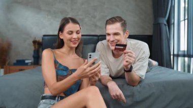 Genç çift, kredi kartıyla ödemek için internet üzerinden ödeme uygulaması ve dijital cüzdan kullanarak yatak odasında oturuyor. Mobil internet üzerinden alışveriş ve modern alımlar. Düzenle