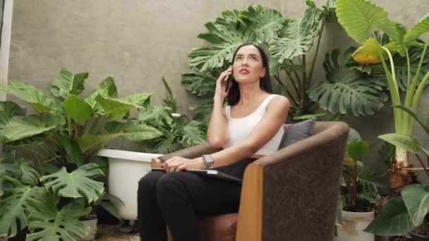 现代的年轻女子在电话中交谈 远程工作或在一个简约的建筑混凝土风格的夏天奇异的植物花园与热带植物的孤独中放松 — 图库视频影像