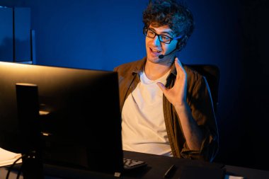 Genç yaratıcı iş adamı, bilgisayar ekranında dinleyicilerle iletişim kuruyor ya da takım üyesiyle görüşüyor. Neon Light Modern Ev Ofisi 'nde kulaklık takıyor. Gece vakti çalışma kavramı. Toplayıcı.