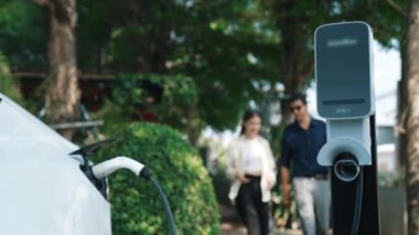 Yavaş çekim çift, ilkbaharda açık yeşil şehir kafesindeki şarj istasyonundan elektrikli araba aküsünü şarj ediyor. Sürdürülebilir çevre dostu seyahat yaşam tarzı için şarj edilebilir EV araba