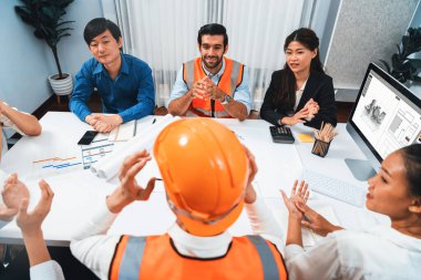 Çeşitli inşaat mühendisi ve müvekkil grupları, mimari proje üzerinde başarılı bir anlaşma sağlandıktan sonra heyecanlanıp alkışlanıyorlar, inşaat planını gözden geçiriyorlar ve toplantı masasında planlarını gözden geçiriyorlar.