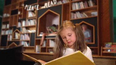 Genç, zeki, beyaz bir kız kütüphanede otururken kitap okuyor. Zeki çocuk öğrenme, okuma, kütüphanede kitap açma. Etkileyici çocuk geçmişi bulanık sayfayı çeviriyor. Etkinlik.