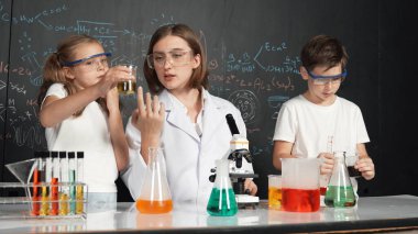 Öğretmen tavsiye verirken beyaz çocuk kimyasal sıvıyı karıştırıyor. Profesyonel eğitmen labaratuar kıyafeti giymiş, renkli solüsyonla dolu deney kabıyla sofrada çeşitli öğrencileri arıyor. Etkinlik.
