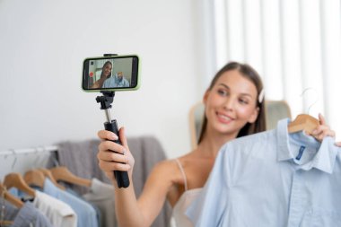 Sosyal medya için moda videosu hazırlayan genç bir kadın. Blogcu kameraya selfie çubuğu ve ışık halkası kullanarak gülümserken, aynı zamanda ikna edici online kıyafetlerin izleyicilere veya takipçilere satılmasını sağlıyor. Açılıyor