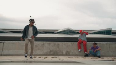 Yakışıklı break dansçı, Asyalı arkadaşının tezahürat yaptığı serbest stil hareketinden hoşlanır. Hippi kulaklıktan müzik dinler ve kameraya bakar. Sokak dansı. Açık hava sporu 2024. Yumuşak bir şekilde..