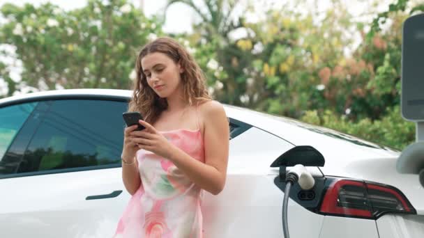 若い女性は 自然の中で公共Evカー充電ステーションで電気を支払うためにスマートフォンを使用しています Ev車両による近代的な環境と持続可能な自動車輸送ライフスタイル ファスティオディオス — ストック動画