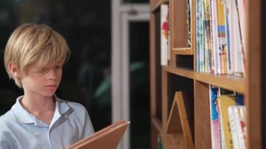 Genç, zeki, beyaz bir çocuk kitap rafından kitap seçiyor ya da okuyor. Zeki çocuk öğrenme, okuma, kütüphanede kitap açma. Çekici çocuk sayfayı çeviriyor. Bir yığın kitap. Etkinlik.