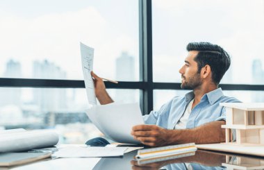 Modern ofisteki masanın üzerinde, yetenekli beyaz mimar mühendisi planları tutuyor. Tasarım ve ev modeli arasında karşılaştırıldığında akıllı bir iç mimar. Proje, plan, inşaat, proje. İzleme.