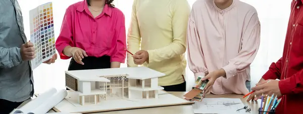 Cropped image of professional architect team brainstorm about house color selection while architect writes on blueprint document at table placed with blueprint and house model. Variegated.