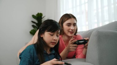 Annem dizüstü bilgisayar kullanarak kanepeye uzanırken, mutlu kız oyun oynuyor ve oturuyor. Beyaz bir ebeveyn ve çekici bir kız, elinde joystick ile birlikte vakit geçirir ve maç kazanmaya odaklanırlar. Pedagoji.