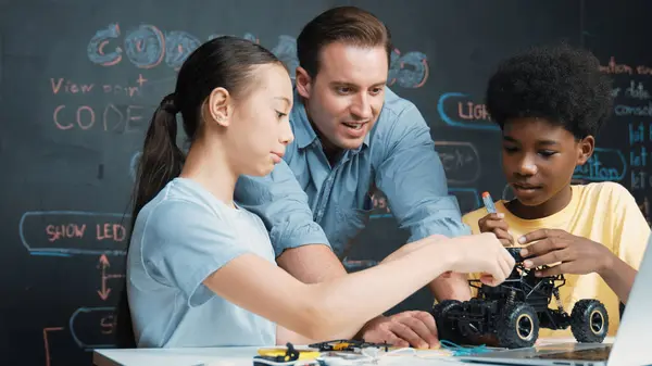 白人老师看着笔记本电脑屏幕 同时用编写的代码在黑板上解释汽车模型系统 快乐多样的学生学习机器人的构造 — 图库照片