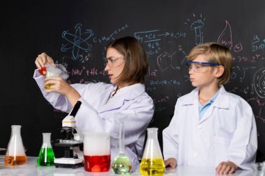 Laboratuvardaki yetenekli öğretmen ve öğrenci laboratuvar önlüğü giyer ve STEM sınıfında kimya bilimi üzerine deney yaparlar. Öğretmen kırmızı sıvıyı sarı şişeye döktü. Masa bir sürü matara koydu. Etkinlik.