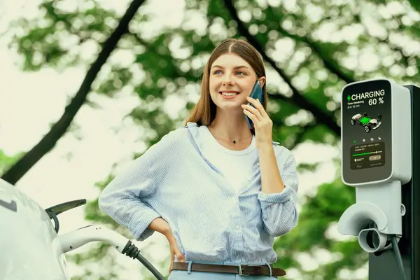 国立公園や夏の森での休暇旅行中にEv充電ステーションから電気自動車バッテリーを充電するためにスマートフォンオンラインバンキングアプリケーションを使用している若い女性 エクスアルト — ストック写真