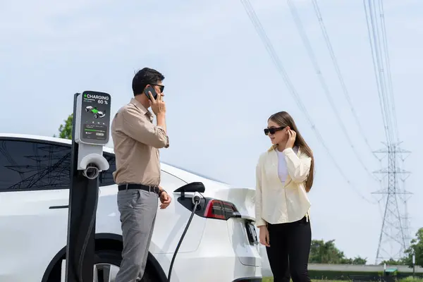 Couple pay for electricity with smartphone while recharge EV car battery at charging station connected to power grid tower electrical as electrical industry for eco friendly car utilization.Expedient