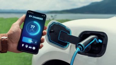 Akıllı telefon ekran akü durumu arayüzünü elinde tutan iş adamı akıllı EV mobil uygulama ile EV otomobili elektrik şarj istasyonundan doğal göl arka planı ile elektrik. İnceleyin.