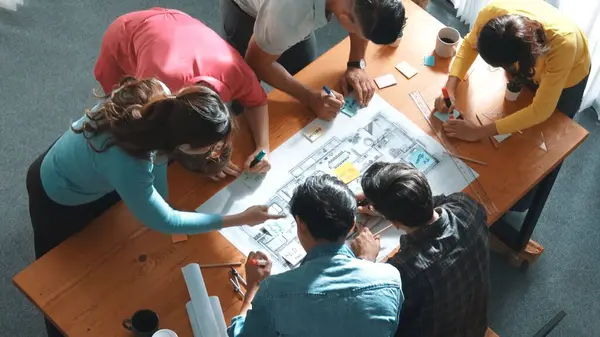 精明能干的土木工程师坐在餐桌前 手握蓝图 建筑设备和五彩缤纷的便条 一边工作一边书写蓝图的最高视图 多样化的团队参加会议 专题讨论会 — 图库照片
