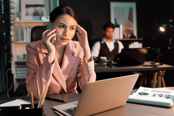Focusing woman calling to customer working at front desk view while waiting email business project report on laptop at late night time with blurry man coworker background at modern office. Postulate.