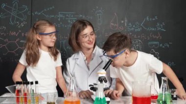 Öğretmen tavsiye verirken beyaz çocuk kimyasal sıvıyı karıştırıyor. Profesyonel eğitmen labaratuar kıyafeti giymiş, renkli solüsyonla dolu deney kabıyla sofrada çeşitli öğrencileri arıyor. Etkinlik.