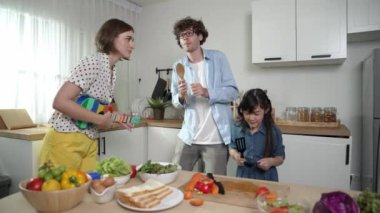 Beyaz yetenekli baba, anne ve Asyalı kız birlikte dans ederken kahvaltı hazırlıyorlar. Modern mutfakta sebze hazırlarken ukulele çalan yetenekli bir anne. Sağlıklı gıda konsepti. Pedagoji.