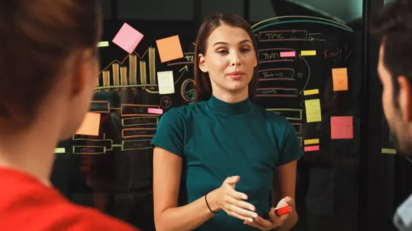 Skilled executive manager explain business idea to diverse investor team. Businesspeople listening female leader present marketing strategy while using sticky notes and mind mapping. Tracery