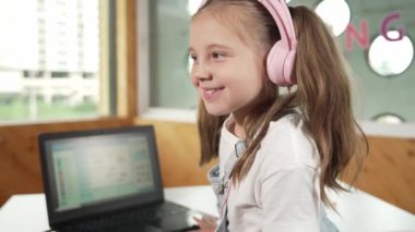 Masaya dizüstü bilgisayarı yerleştirip kameraya el sallayan gülümseyen kız. BTMM sınıfında dizüstü bilgisayar programlama ya da kodlama programı yaparken kulaklık takan bir çocuk gülümsüyor. Etkinlik.