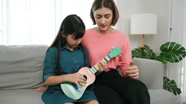 ハッピーガールはウクレレを演奏しながら ママは自宅でアコースティック音楽を教え 説明します 楽器について学ぶかわいい子供 ハッピーな白人の母親と少女は一緒に時間を過ごしています ペダゴロジー — ストック写真