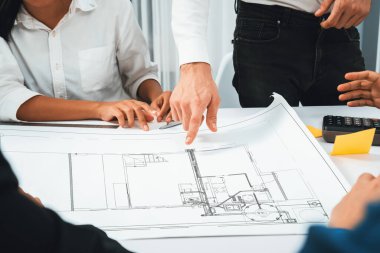 Mimari proje üzerinde birlikte çalışan çeşitli inşaat mühendisi ve müvekkil grupları, inşaat planını gözden geçirip toplantı masasında plan oluşturuyorlar. Tedbirli