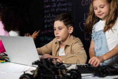 STEM sınıfında sınıf öğrencisi robot araba kodlamayı öğreniyor. Kahverengi gömlekli çocuk ve beyaz önlüklü kız dizüstü bilgisayar izleyip tartışıyorlar. Diğer liseli kızlar komik oyunlar oynuyorlar. Etkinlik.