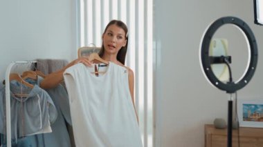 Genç sosyal medyum kadın moda videosu yapıyor. Blogcu kameraya selfie çubuğu ve ışık halkası kullanarak gülümserken, aynı zamanda ikna edici çevrimiçi kıyafetlerin izleyicilere veya takipçilere vlog satmasını sağlıyor. Düzenle
