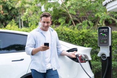 Modern çevre dostu adam EV şarj istasyonundan elektrikli aracı şarj ediyor. Evde şarj edilen pilleri optimize etmek için enerji kullanımını takip etmek için yenilikçi EV teknolojisi kullanımı. Eşzamanlama