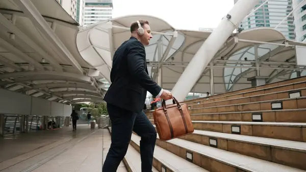 Smart caucasian businessman walking up stairs surrounded by urban view. Back view of happy ambitious project manager listen relaxing music from headphone and move to song while climb stairs. Urbane.