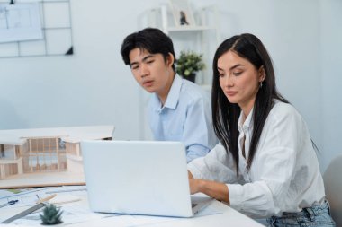 Profesyonel Asyalı erkek mimar ev modelinin uzunluğunu ölçmek için cetvel kullanırken, beyaz tenli genç meslektaşı ev modeliyle toplantı masasındaki verileri analiz etmek için dizüstü bilgisayar kullanıyor. Tertemiz..