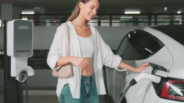 Yavaş hareket eden kadın mutlu ve EV elektrikli arabasıyla şehir merkezindeki Shopp Center otoparkına doğru gülümsüyor. Elektrikli taşıt iç organları için EV şarj prizi taşıyor.