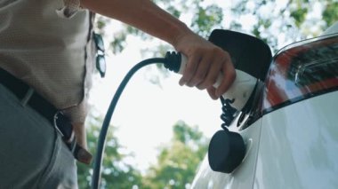 Yavaş çekim adamı, yaz mevsiminde açık yeşil yay yeşilliğindeki şarj istasyonundan elektrikli araba aküsünü şarj etmek için EV şarj aleti taktı. Yeniden şarj edilebilir elektrikli araç için sürdürülebilir enerji
