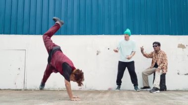 Genç Asyalı dansçı hip hop tarzında dans ediyor ve çok kültürlü arkadaşları duvarın arkasında tezahürat yapıyor. Çekici yakışıklı adam mavi arka planda kollarını uzatıyor. Açık hava sporu 2024 hiphop..