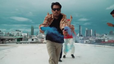 Bir grup çok kültürlü hippi çatıda işaret fişeği çalıyor. Zenci dumanı tutan çekici bir sokak dansçısı hip-hop müzik eşliğinde break dans yapıyor. Açık hava sporu 2024. Sokak dansı. hiphop.