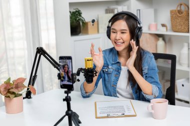 Asyalı sunucu canlı yayında canlı yayında canlı yayında sosyal medyada kulaklık takarak konuşuyor. Dinleyicileri modern stüdyoda koçluk yaparak selamlıyor. Taktik.