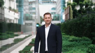 Kafkasyalı mutlu iş adamı, gülümsüyor ve şehir merkezinde kameraya bakıyor. Yeşil şehirde kendine güveni olan yönetici, arka planda bulanık takım elbise giyer. Yeşil çevre. Urbane.