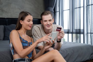 Genç çift, kredi kartıyla ödemek için internet üzerinden ödeme uygulaması ve dijital cüzdan kullanarak yatak odasında oturuyor. Mobil internet üzerinden alışveriş ve modern alımlar. Açılıyor
