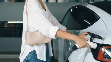 Genç hareketli kadın, EV elektrikli arabasıyla şehir merkezindeki alışveriş merkezinin otoparkına gidiyor. Sürdürülebilirlik yaşam tarzını yeşil, temiz, yeniden şarj edilebilir elektrikli araç iç organlarıyla şarj ediyor.