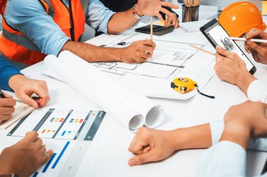 Mimari proje üzerinde birlikte çalışan çeşitli inşaat mühendisi ve müvekkil grupları, inşaat planını gözden geçirip toplantı masasında plan oluşturuyorlar. Tedbirli