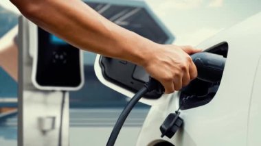 Adam elektrikli araca akıllı EV şarj cihazı takıyor ve EV şarj istasyonundan modern tasarım binası ya da arka planda evi olan şarj oluyor. Geleceksel sürdürülebilir güç ve EV araba kullanımı. İnceleyin