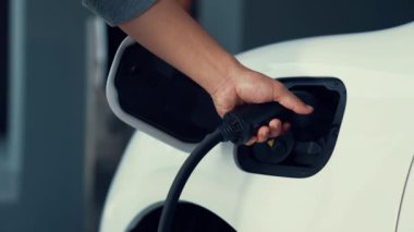 Adam elektrikli arabasını akıllı ev şarj istasyonundaki EV şarj cihazıyla şarj ediyor. Fütürist dijital batarya durumu hologramı. Sürdürülebilir güç için EV araba ve ev enerjisi altyapısı. İnceleyin