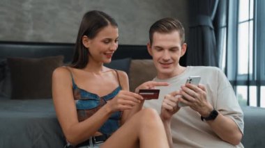 Genç çift, kredi kartıyla ödemek için internet üzerinden ödeme uygulaması ve dijital cüzdan kullanarak yatak odasında oturuyor. Mobil internet üzerinden alışveriş ve modern alımlar. Düzenle