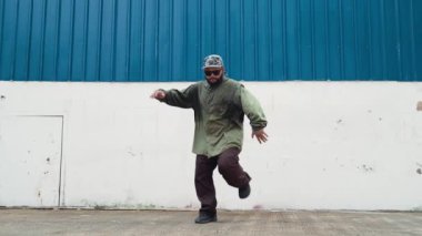 Sokak ortasında mavi duvarlı hippi dansçı ayak basamağı. Asyalı break dansçı kollarını açarken sokak dansı yapıyor. Hip hop koreografı enerjik bir dans sergiliyor. Açık hava sporu 2024 hiphop..