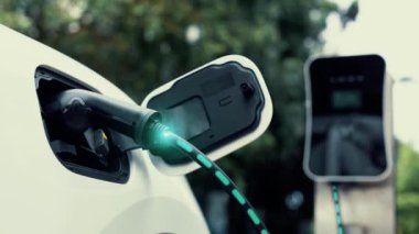 Elektrikli araba, doğal parktaki temiz enerji şarj istasyonundan gelen fütürist akıllı EV şarj cihazının dijital batarya durumu hologramıyla şarj oluyor. Gelecekçi çevre dostu EV araba. İnceleyin