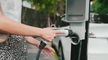 Şehir merkezindeki EV şarj istasyonunda elektrikli araba aküsünü yavaş çekimde şarj eden kadın elektrikli aracın iç organları için sürdürülebilir yeşil ve yenilenebilir temiz enerji yaşam tarzına sahip.