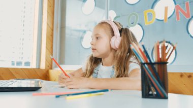 Tatlı akıllı kız sınıf çalışması yapıyor ya da öğretmenini dinlerken internetten öğreniyor. Mutlu öğrenci yazıları, çizimleri, bilgisayar ve renkli kalemlerle kulaklık takarak kağıt üzerinde çalışması. Etkinlik.