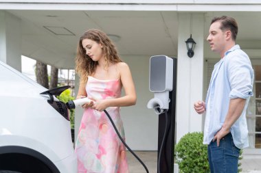 Çevre dostu, mutlu ve sevimli çift EV şarj istasyonundan elektrikli aracını şarj ediyor. EV araba teknolojisi gelecekteki sürdürülebilirlik için kullanılıyor. Eşzamanlama